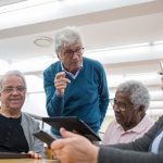 Grupo de idosos participa de uma discussão sobre tecnologia usando tablets e laptops em um ambiente interno.