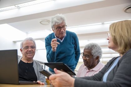 Grupo de idosos participa de uma discussão sobre tecnologia usando tablets e laptops em um ambiente interno.