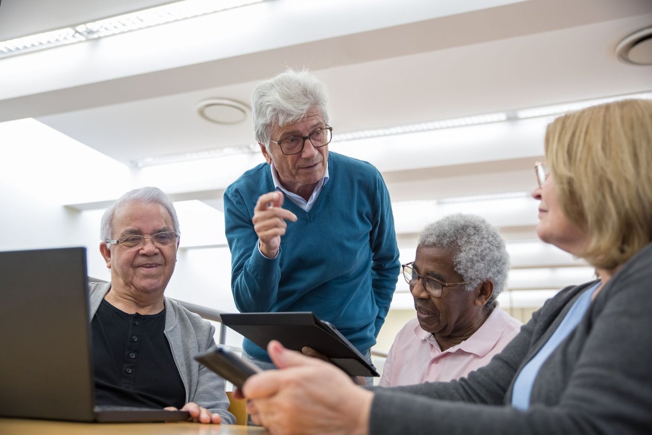Grupo de idosos participa de uma discussão sobre tecnologia usando tablets e laptops em um ambiente interno.