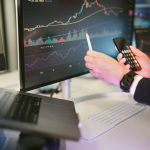 Profissional analisa gráficos financeiros em monitores e consulta um smartphone em um ambiente de trabalho digital.