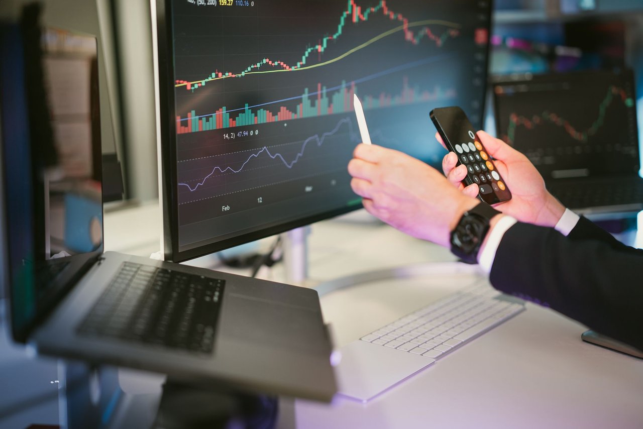 Profissional analisa gráficos financeiros em monitores e consulta um smartphone em um ambiente de trabalho digital.