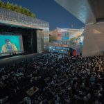 Público reunido ao ar livre durante uma sessão noturna de cinema sob um viaduto.