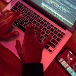 Mãos digitando em um laptop com código na tela, ao lado de um smartphone, em um ambiente interno pouco iluminado