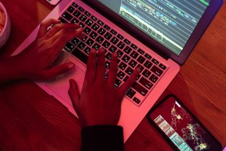 Mãos digitando em um laptop com código na tela, ao lado de um smartphone, em um ambiente interno pouco iluminado