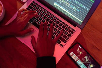 Mãos digitando em um laptop com código na tela, ao lado de um smartphone, em um ambiente interno pouco iluminado