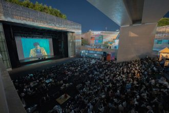 Público reunido ao ar livre durante uma sessão noturna de cinema sob um viaduto.